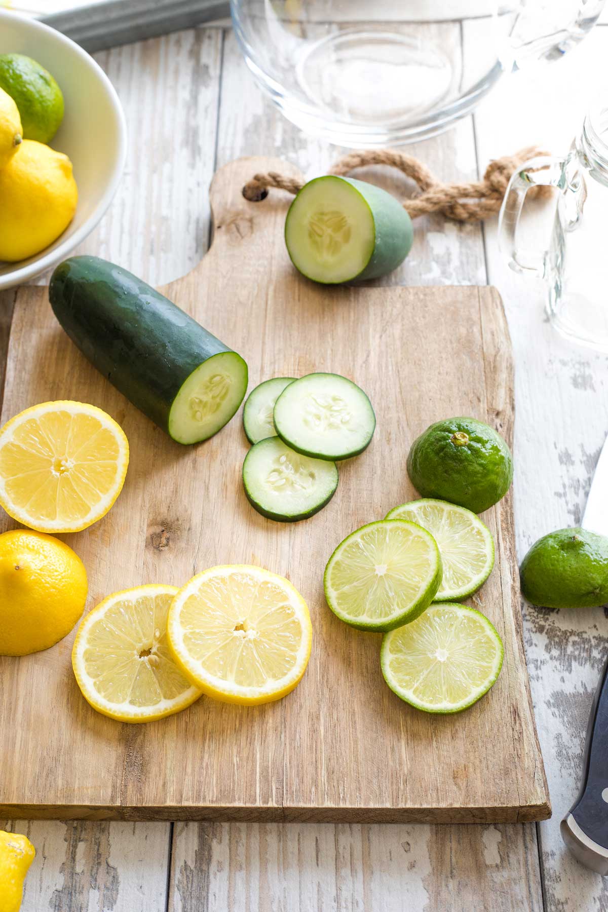 Sliced lemon, lime and cucumber on wooden board with empty water pitcher in background.