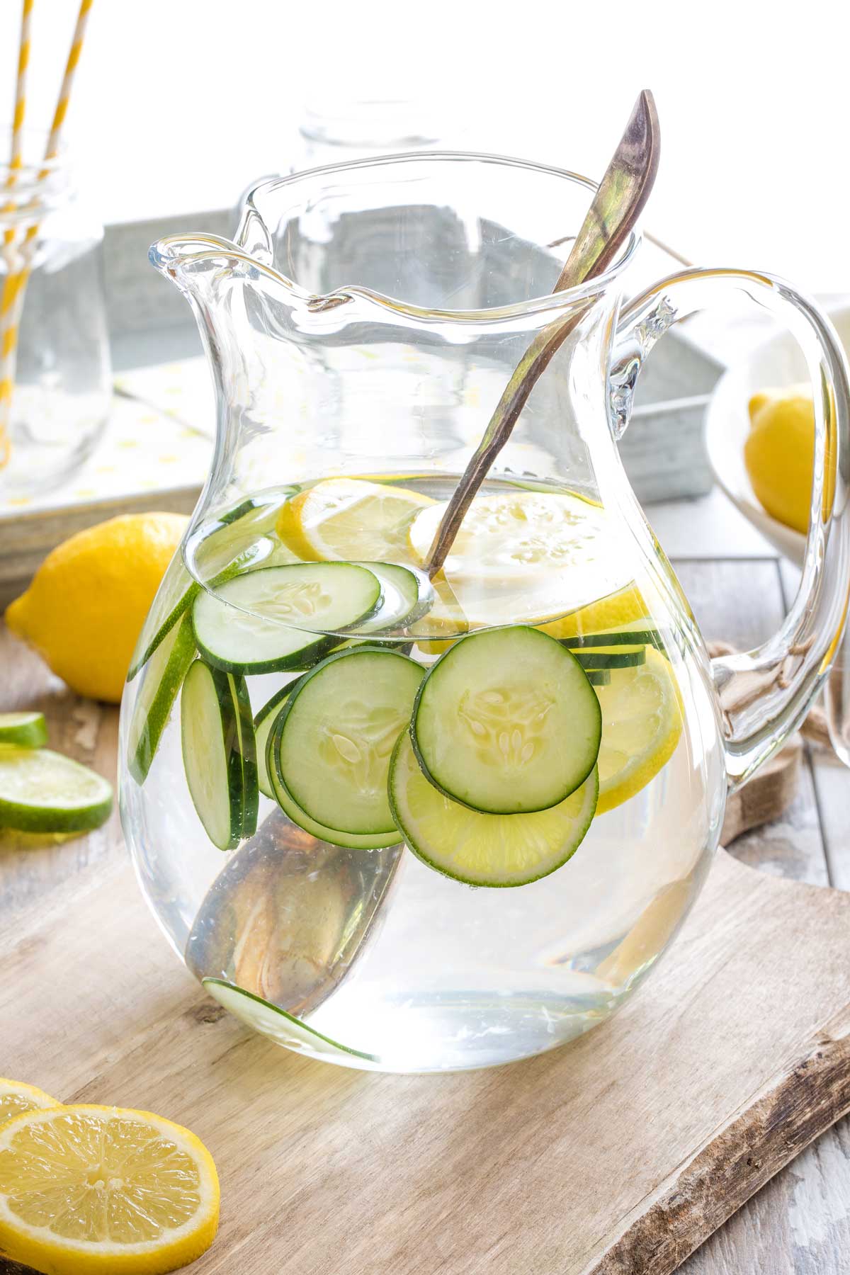 Pitcher of freshly made infused water on cutting board with large antique spoon in it to stir, and additional lemon and lime slices nearby on cutting board.