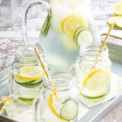 Picnic scene on distressed white wood with full pitcher of water and three glasses on metal serving tray with yellow and green spotted papers, striped paper straws and lots of slices of lemons, limes and cucumbers in water.