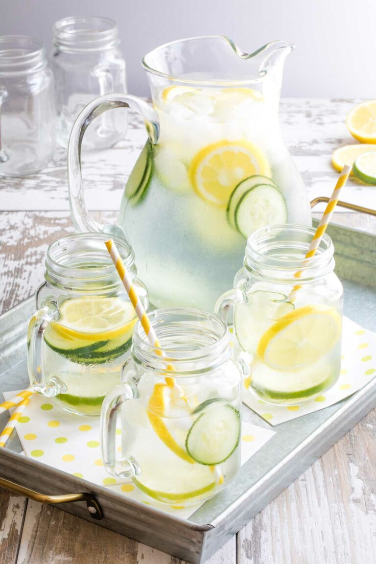 Picnic scene on distressed white wood with full pitcher of water and three glasses on metal serving tray with yellow and green spotted papers, striped paper straws and lots of slices of lemons, limes and cucumbers in water.