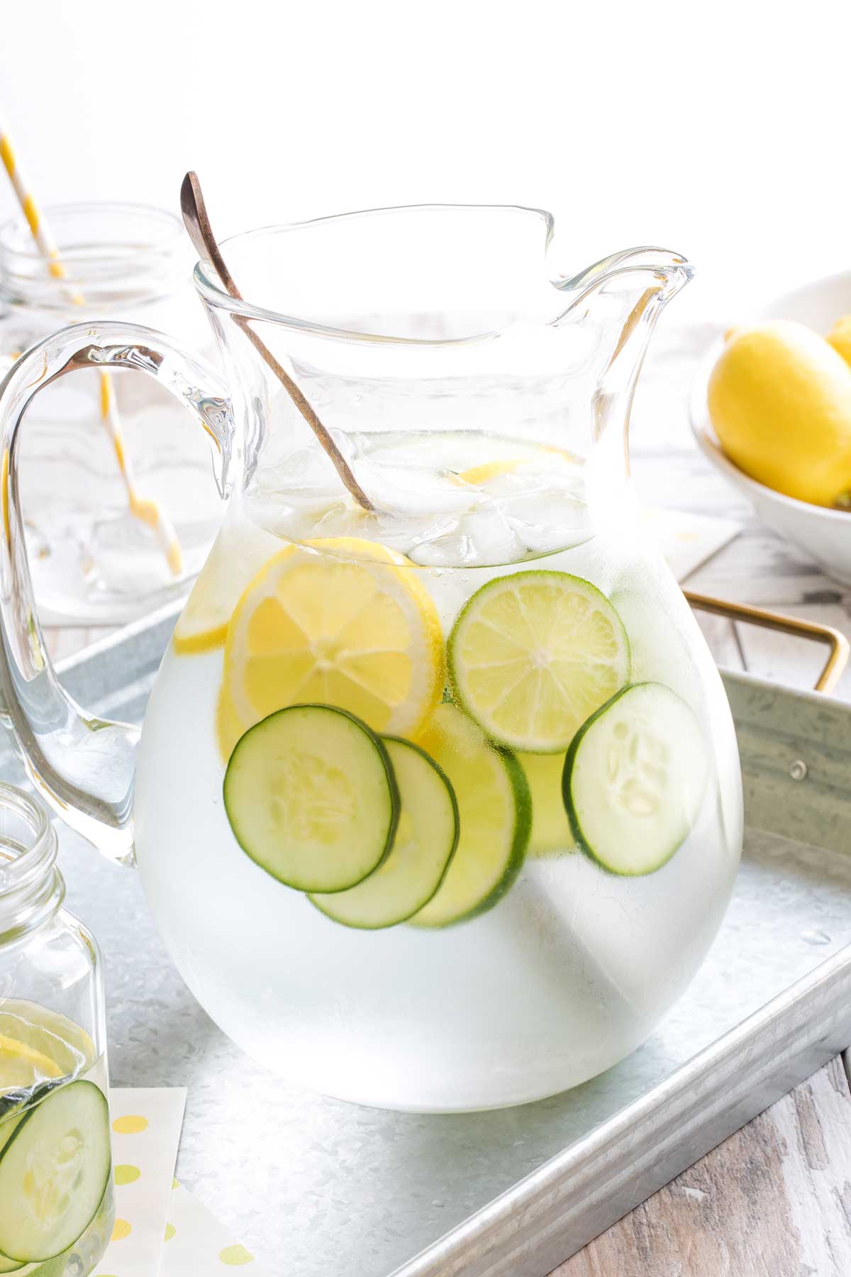 Condensation-covered pitcher of this water recipe, with spoon in it, plus slices of cucumber, lemons and limes and melting ice cubes, sitting on metal picnic tray.