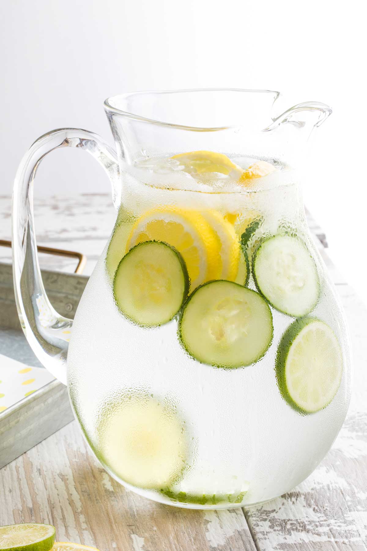 Condensation-covered pitcher of water on distressed white wood, with slice of lemons, limes and cucumber and small ice cubes floating in it.
