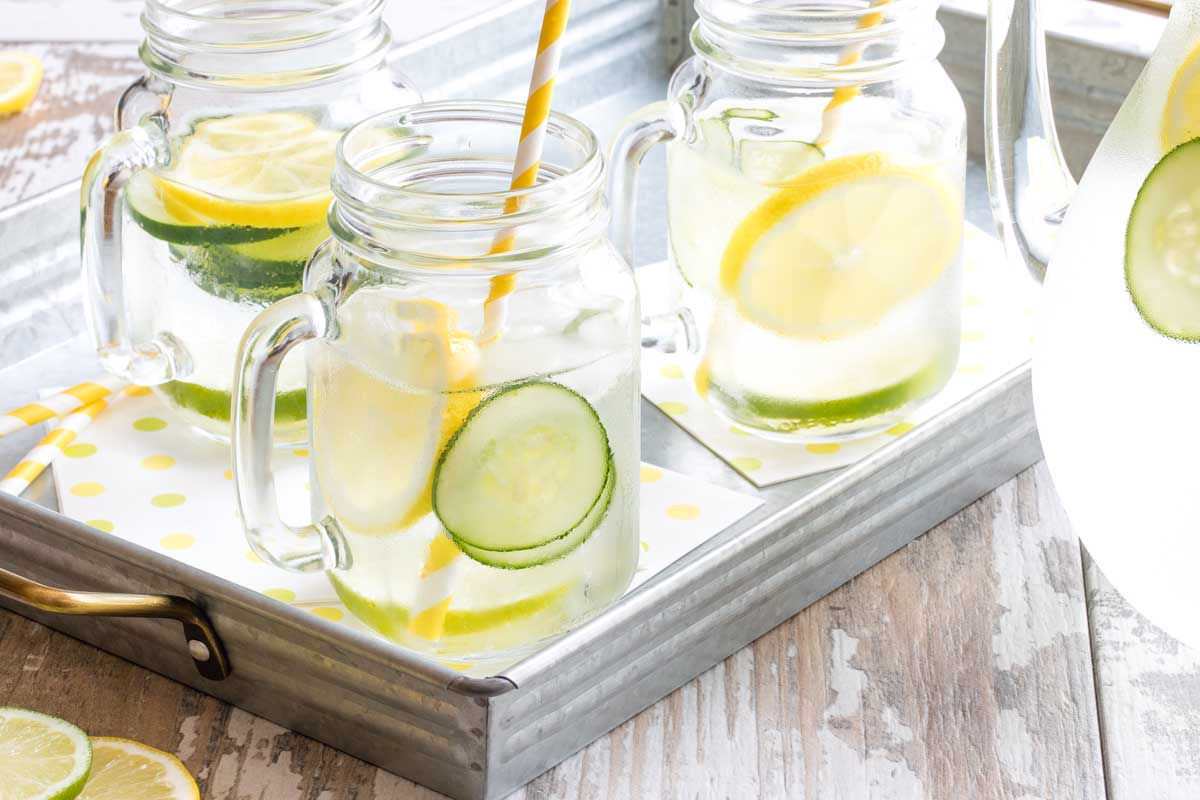 Three Mason jar glasses full of water with yellow striped straws and slices of cucumber, lemon and lime; on metal tray with water pitcher at side.