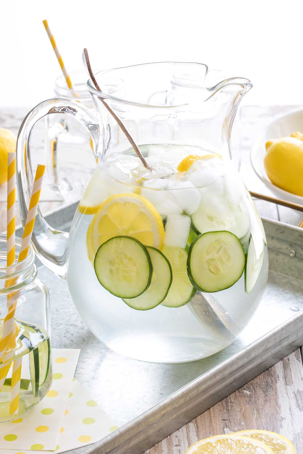 Glass pitcher full of Lemon Lime Cucumber Water and ice cubes, with large antique spoon in it, on metal tray with extra lemons, glasses and straws.