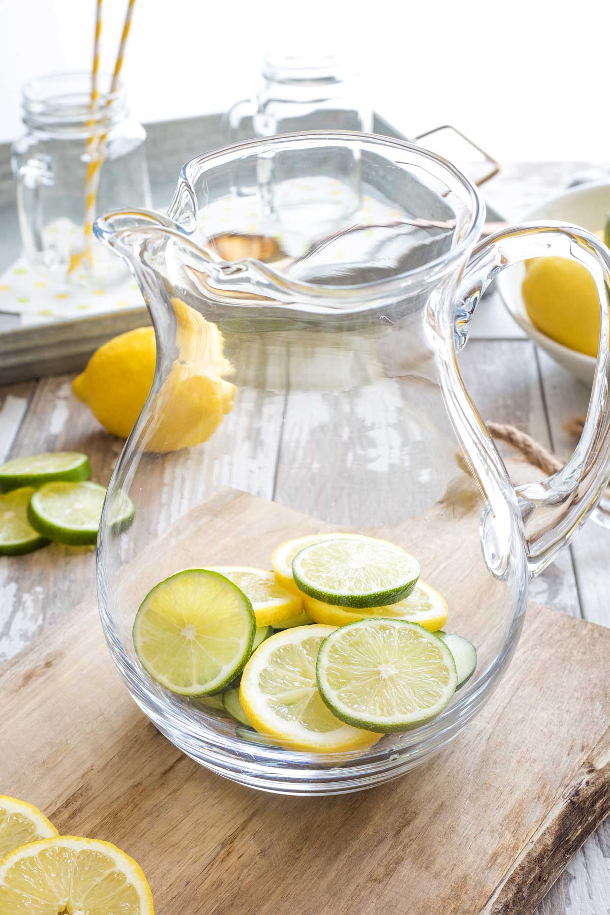 Water pitcher on cutting board, with lemon, lime and cucumber slices in bottom before water is added.