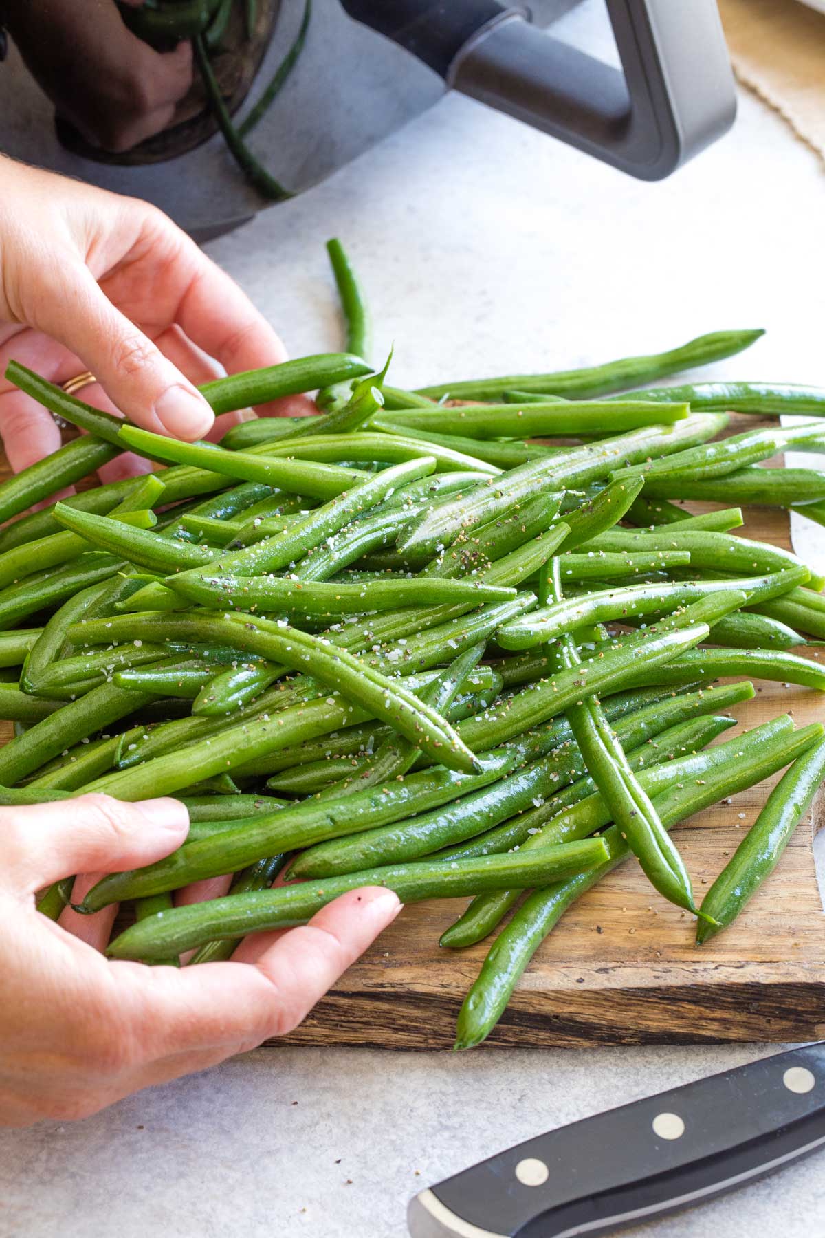 Two hands tossing raw green beans with oil and seasonings, with air fryer waiting behind.
