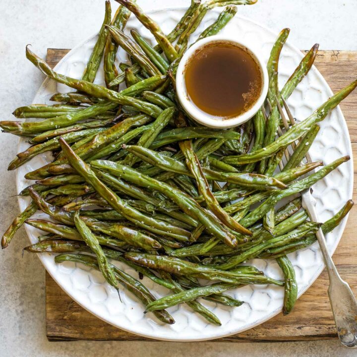Overhead of green beans randomly piled on white platter with little bowl of brown butter sauce and fork entwined in beans.
