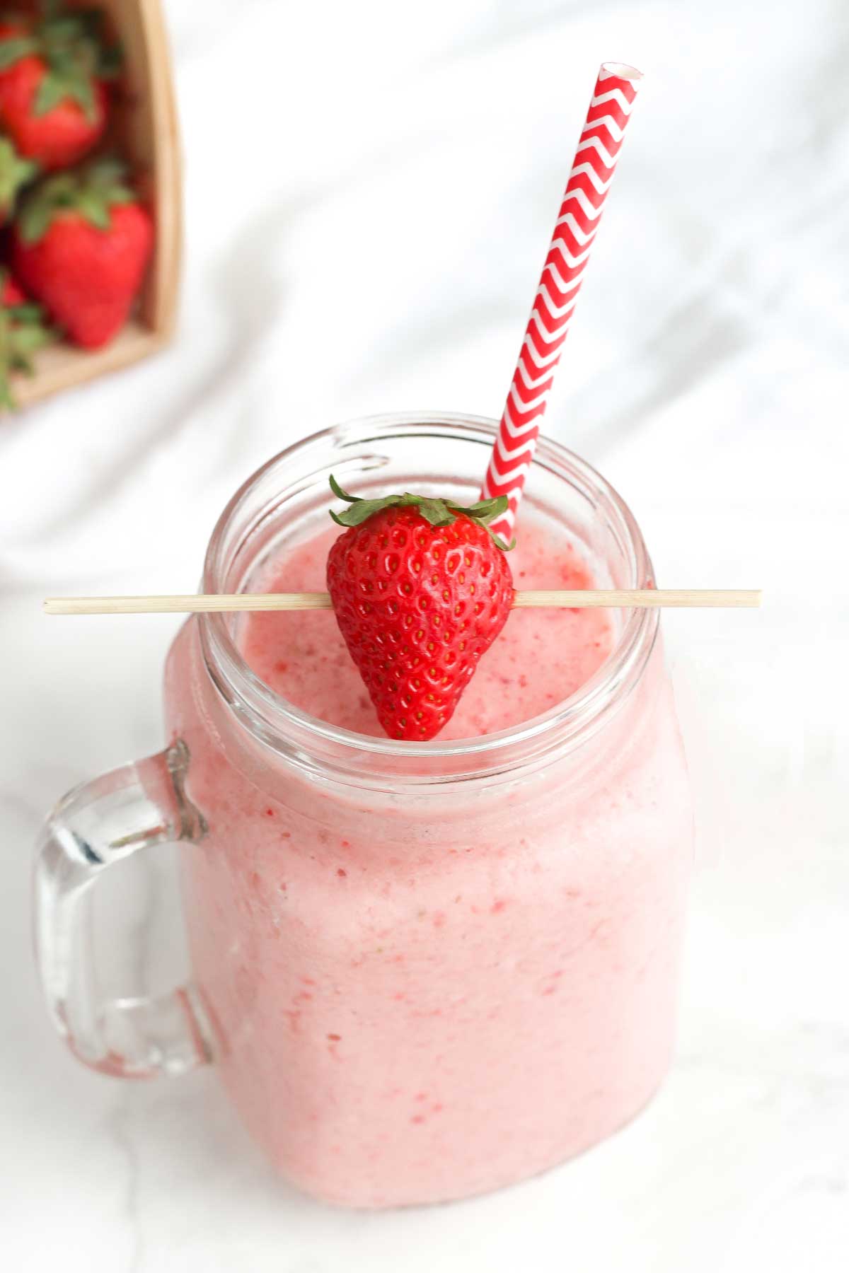 Smoothie in clear, handled, jar-style mug with red and white striped paper straw and a whole strawberry suspended across the top on a wooden pick.
