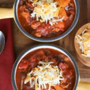 Overhead of two yellow enameled crocks full of chili,. topped with shredded cheese with a little wooden bowl of cheese, a spoon and burgundy napkin alongside.