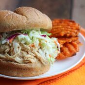 Side closeup of one BBQ chicken sandwich topped with coleslaw on a white plate with a few sweet potato waffle fries alongside.