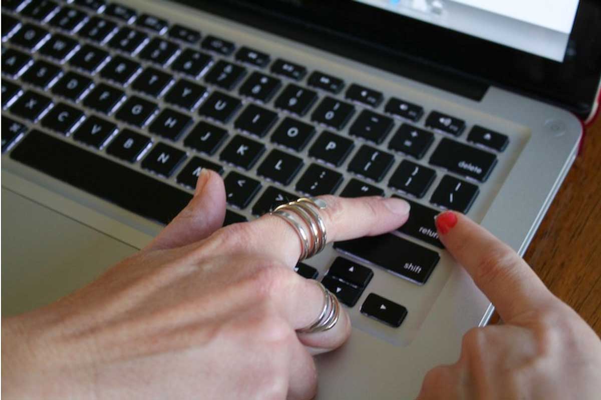 Two different women's hands using index fingers together to hit the return / enter key on a Mac laptop keyboard.