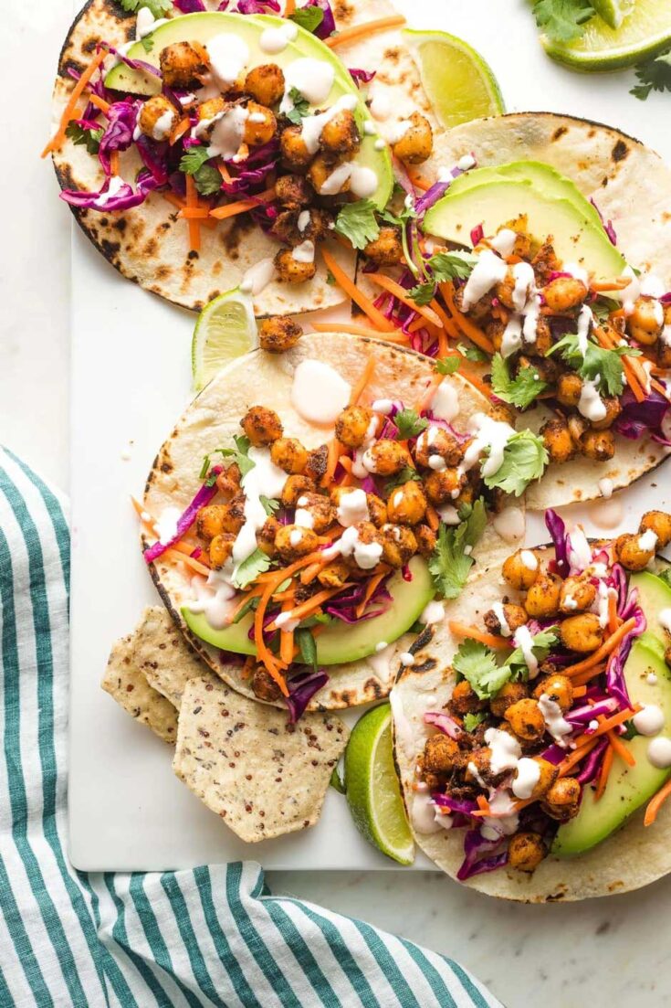 Overhead of 4 tacos open flat and overlapping on white rectangular platter with green and white cloth tucked under.