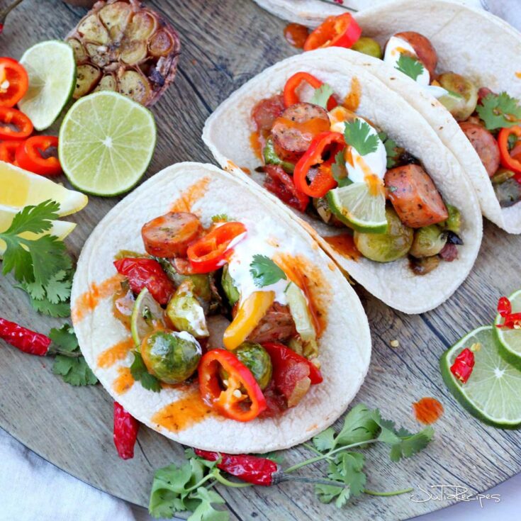 3 tacos in flour tortillas open on round wooden tray with different ingredients surrounding.
