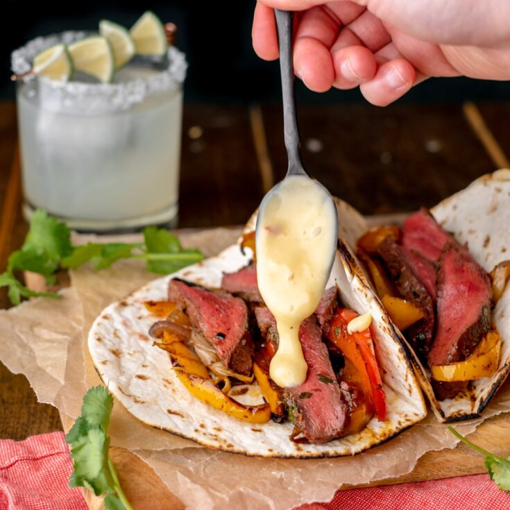 Hand holding spoon to drizzle sauce on one of two tacos with margarita in background.