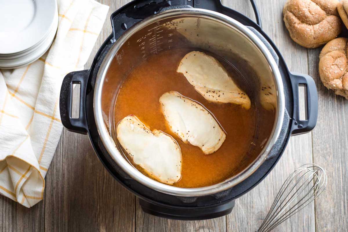3 cooked chicken breasts in orange broth in Instant Pot with buns, whisk, plates and yellow-striped cloth nearby.
