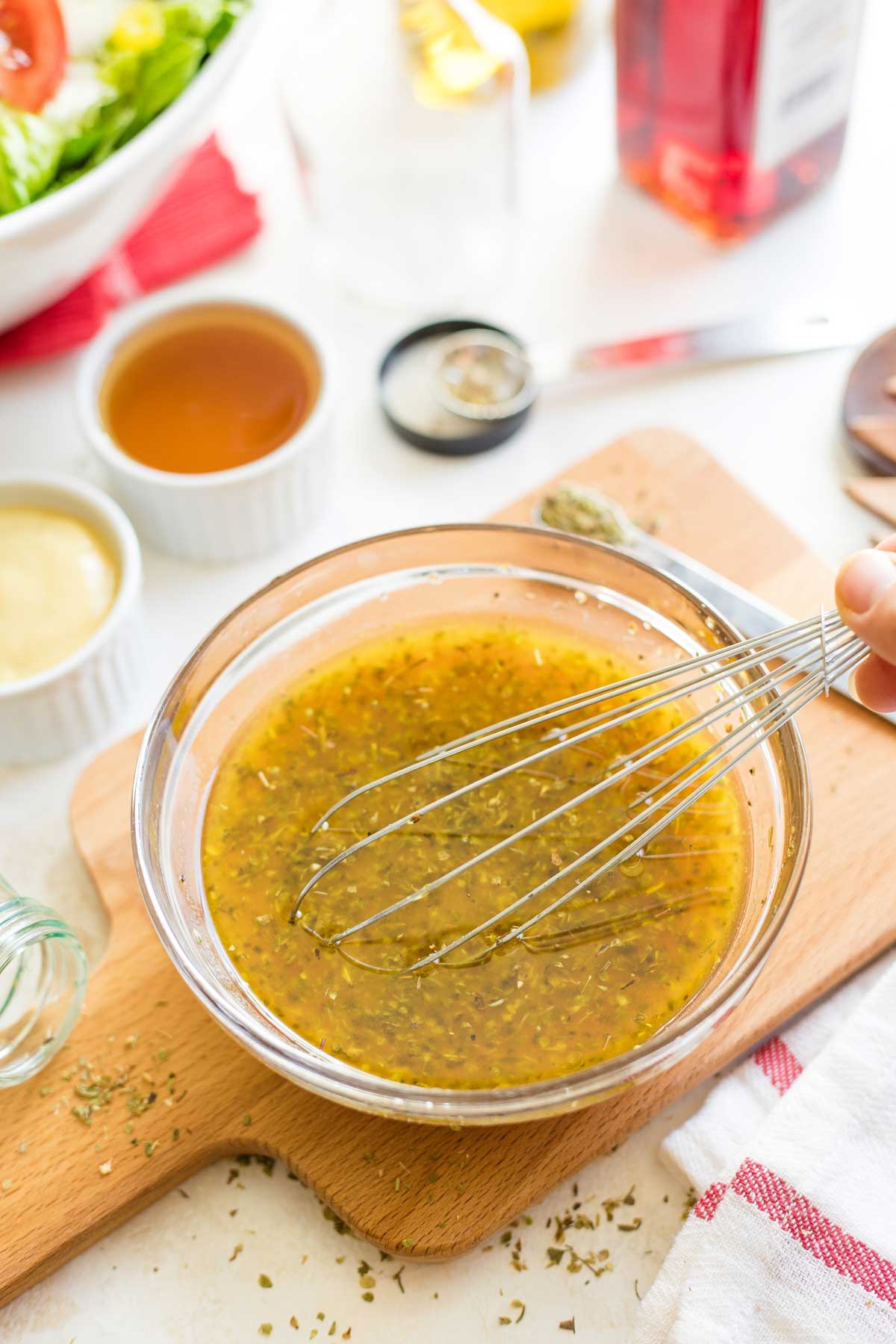 Hand holding whisk in bowl of dressing with ingredients and measuring spoons surrounding.