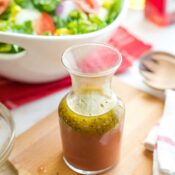 Bottle of vinaigrette on wooden cutting board with Italian salad and bottles of olive oil and red wine vinegar behind.