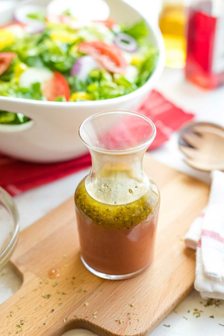 Bottle of vinaigrette on wooden cutting board with Italian salad and bottles of olive oil and red wine vinegar behind.