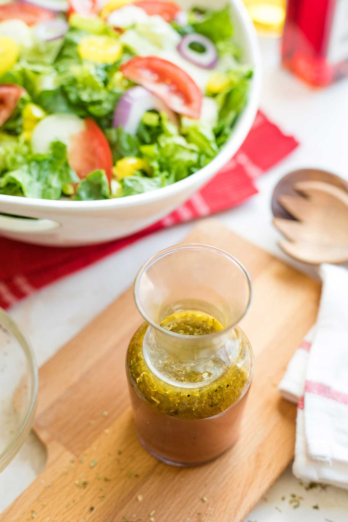 Looking down into bottle of vinaigrette on wooden board, with Italian salad, servers and bottle of red wine vinegar surrounding.