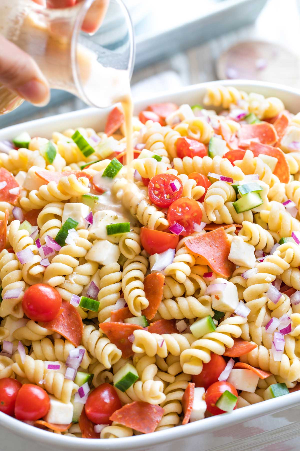 Hand pouring vinaigrette from dressing bottle onto large bowl of Italian pasta salad.