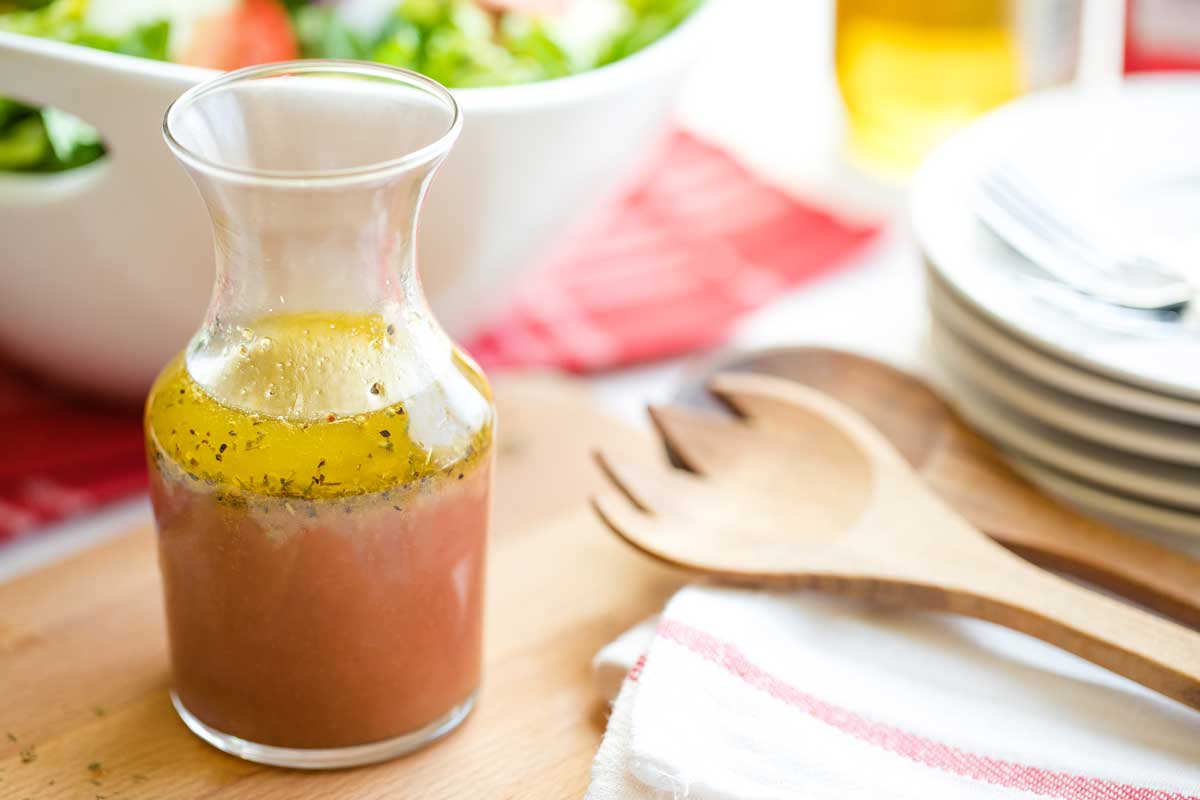Side closeup of bottle of vinaigrette with wooden salad servers, stack of plates and forks and serving bowl of salad greens.