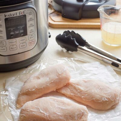 3 pieces of frozen chicken with Instant Pot and lid, tongs and cooking broth.
