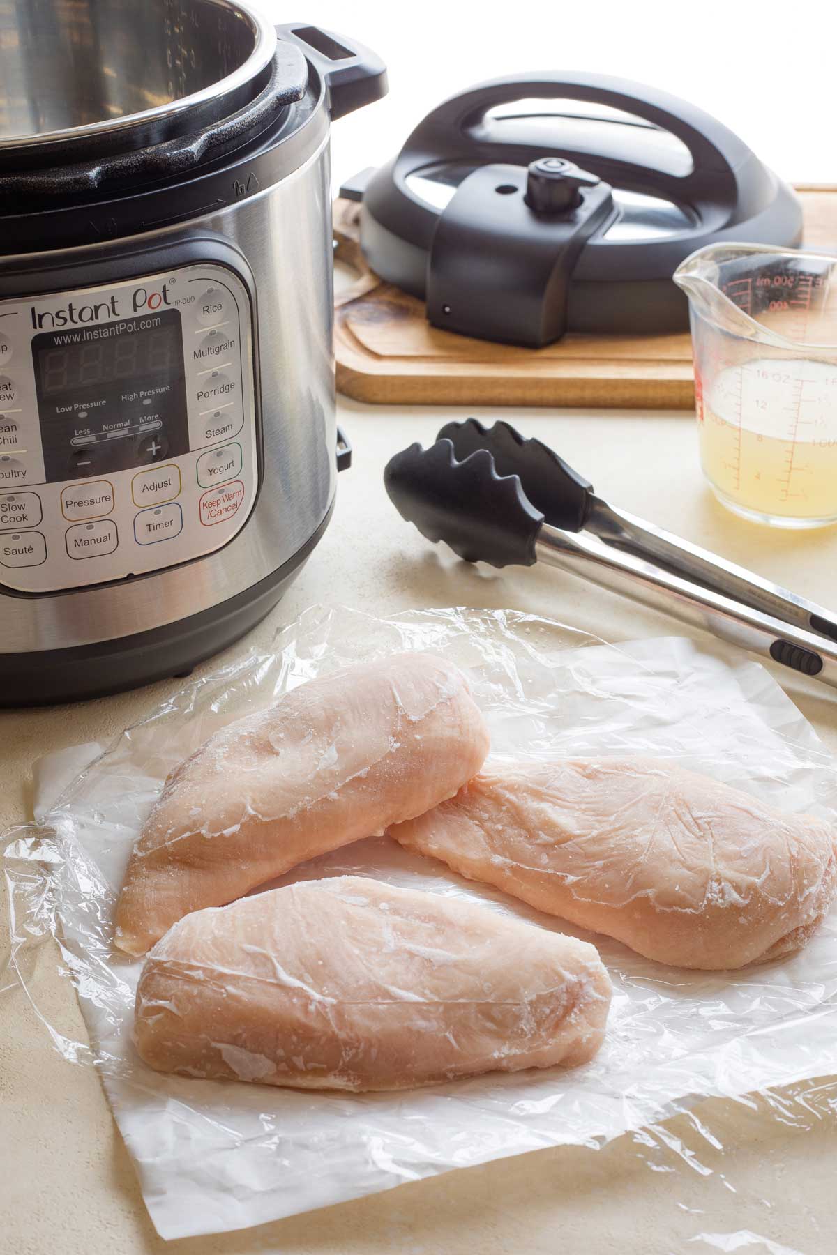 3 frozen chicken breasts on top of plastic wrap and freezer paper, with Instant Pot, tongs and chicken broth behind.