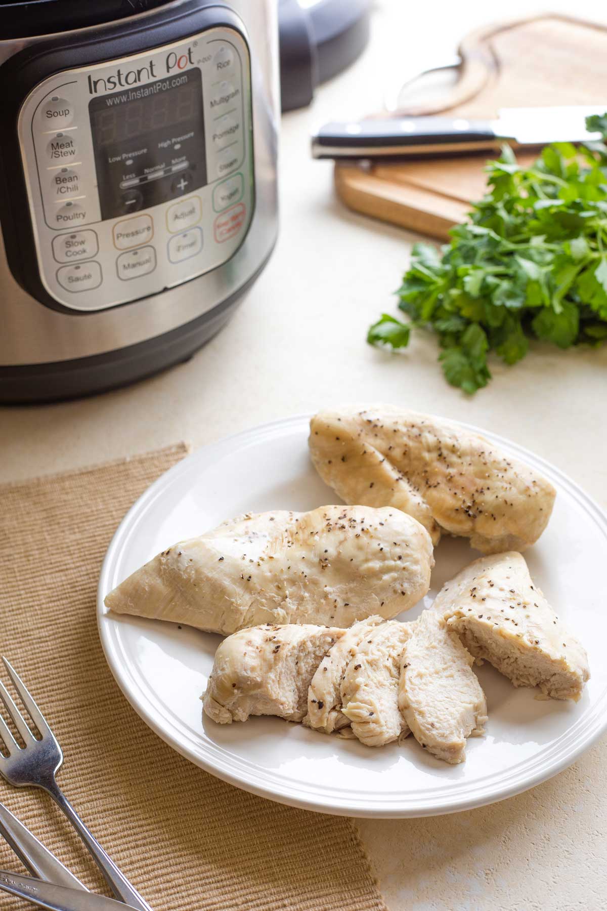 Cooked chicken breasts on white dinner plate (1 sliced) with placemat, forks, Instant Pot and parsley surrounding.