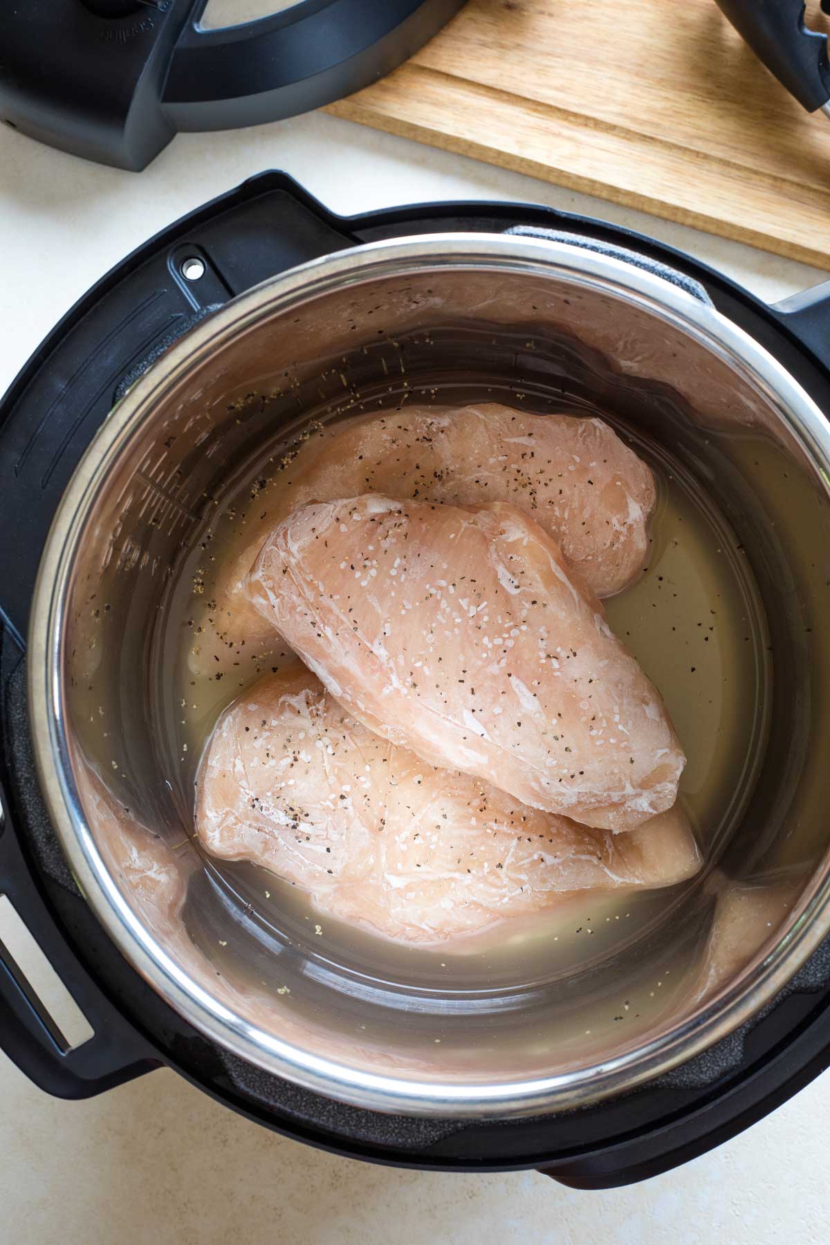 Overhead of 3 frozen chicken breasts in Instant Pot, in broth and sprinkled with salt and pepper - ready to cook.