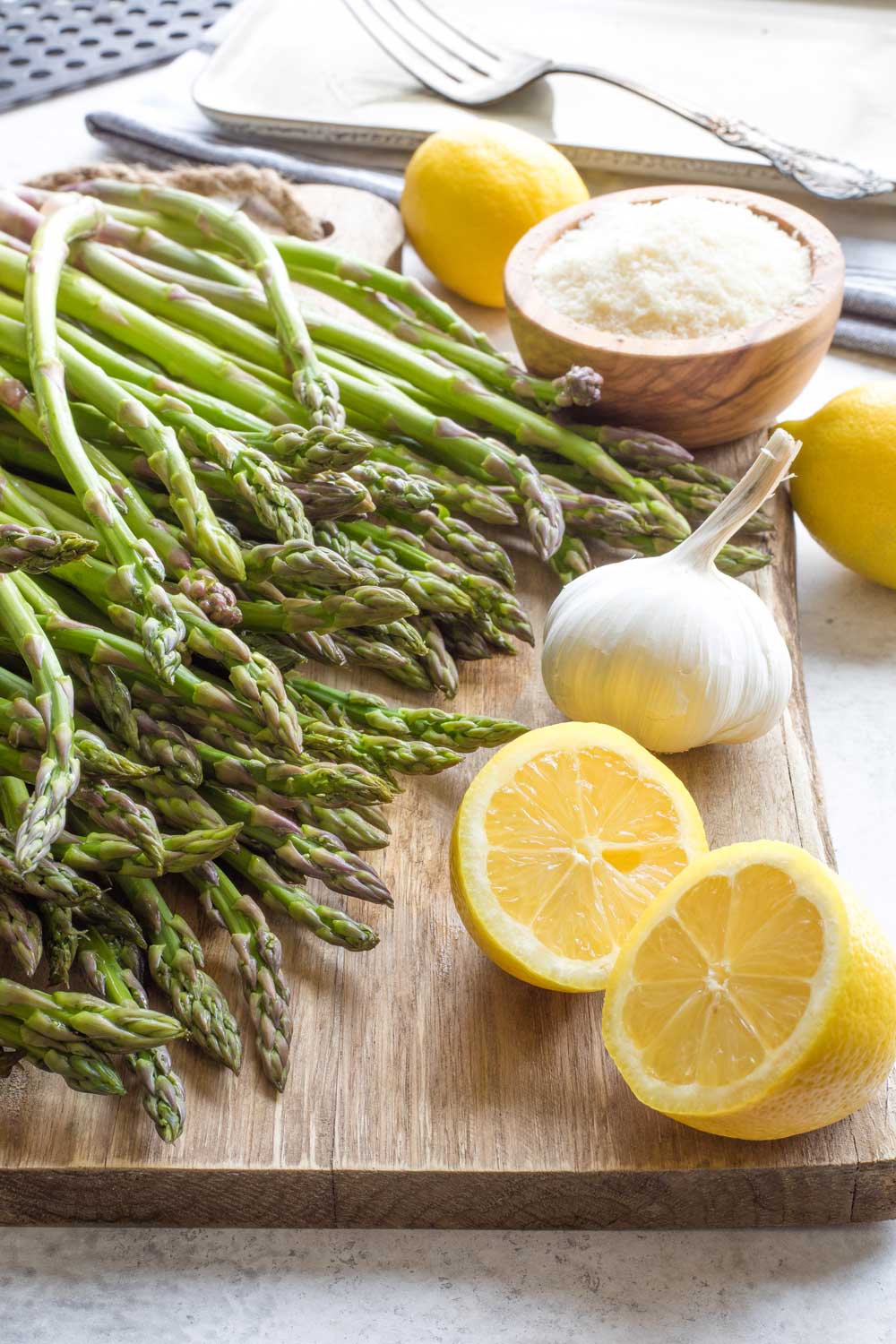 Raw asparagus on cutting board with lemons, head of garlic and bowl of parmesan cheese.