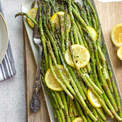 Flatlay of grilled asparagus presented with lemon slices on rectangular platter with antique fork on wood board.