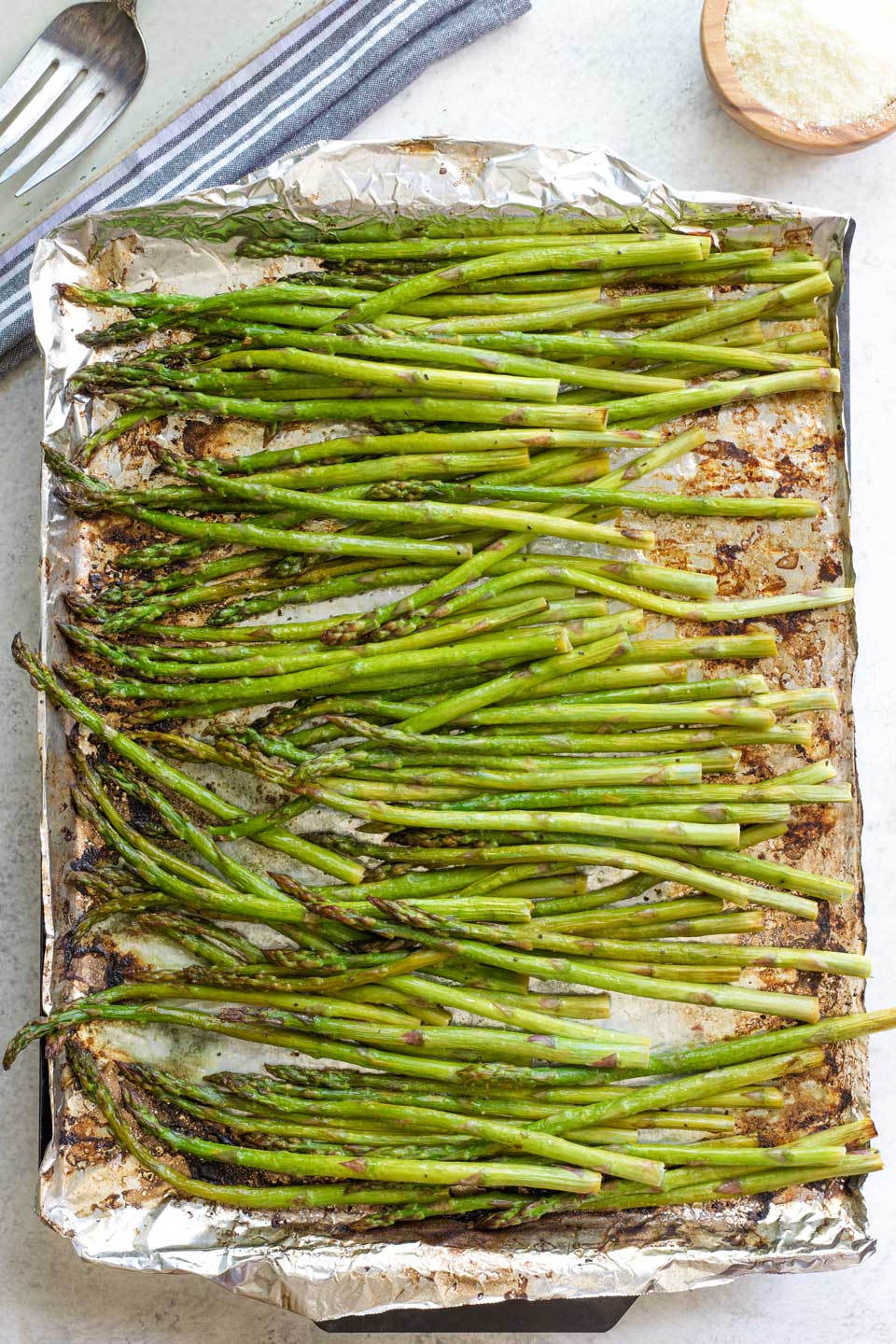 Overhead of asparagus still on pan after grilling, bowl of parmesan and serving platter nearby.