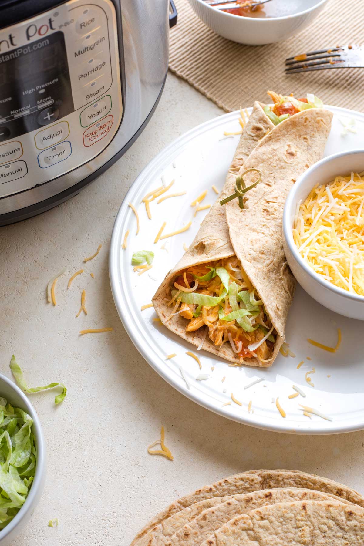 Soft chicken taco rolled up on plate nest to bowl of cheese with tortillas and Instant Pot surrounding.