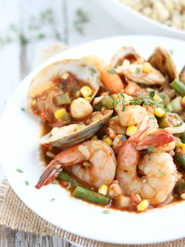 Seafood stew in rimmed bowl on burlap placemat with fresh thyme and rice in background.