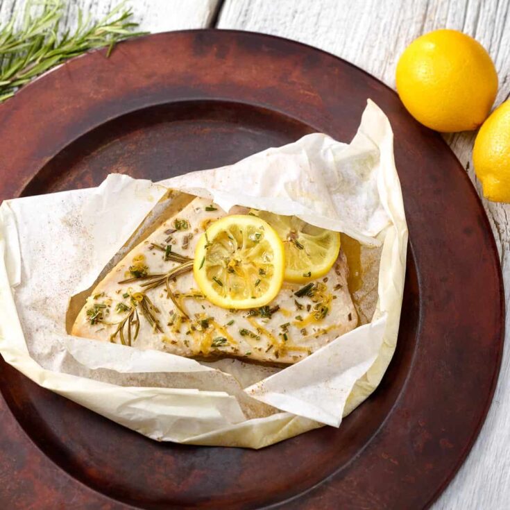 Overhead of one parchment packet opened on bronze charger to reveal lemon-topped fish inside.