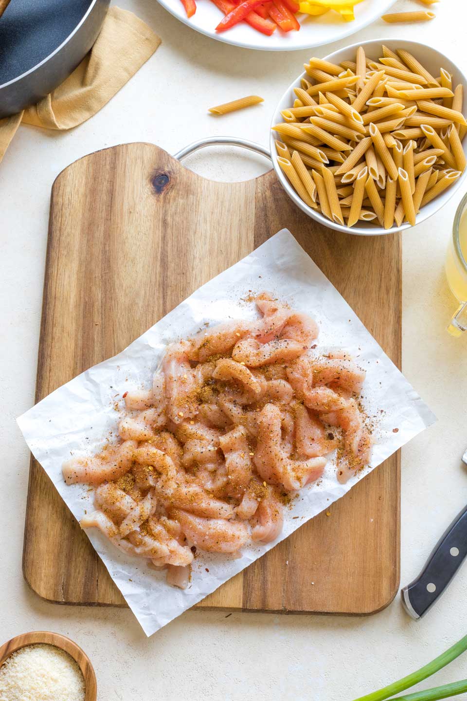 Overhead of chicken strips tossed with jerk seasonings on wooden board with bowl of uncooked pasta.