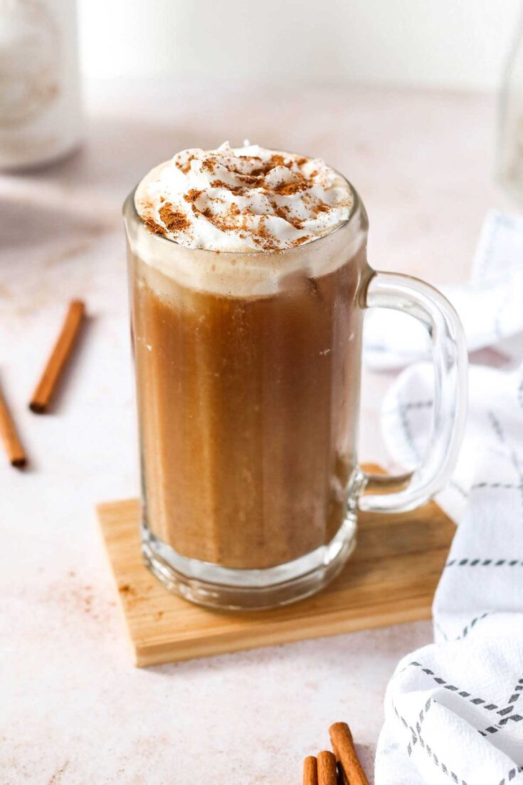 Drink in tall glass mug, topped with whipped cream and cinnamon, surrounded by whole cinnamon sticks.