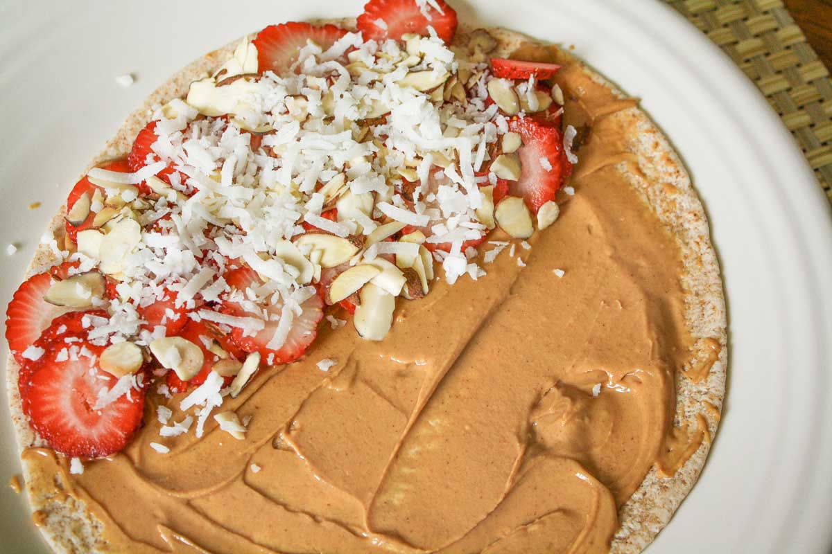 Closeup of tortilla open on plate, smeared with peanut butter, and half topped with strawberries, almonds and coconut.
