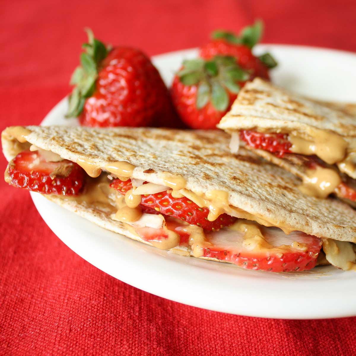 Side view of tortilla wedge so you can see the berries, nuts and peanut butter inside; red background with 3 strawberries.