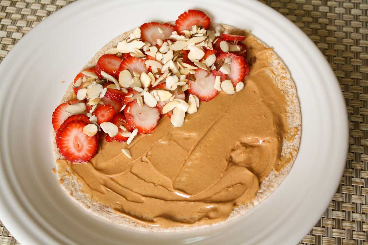 Tortilla on white plate, spread with peanut butter, half topped with strawberry and almond slices.