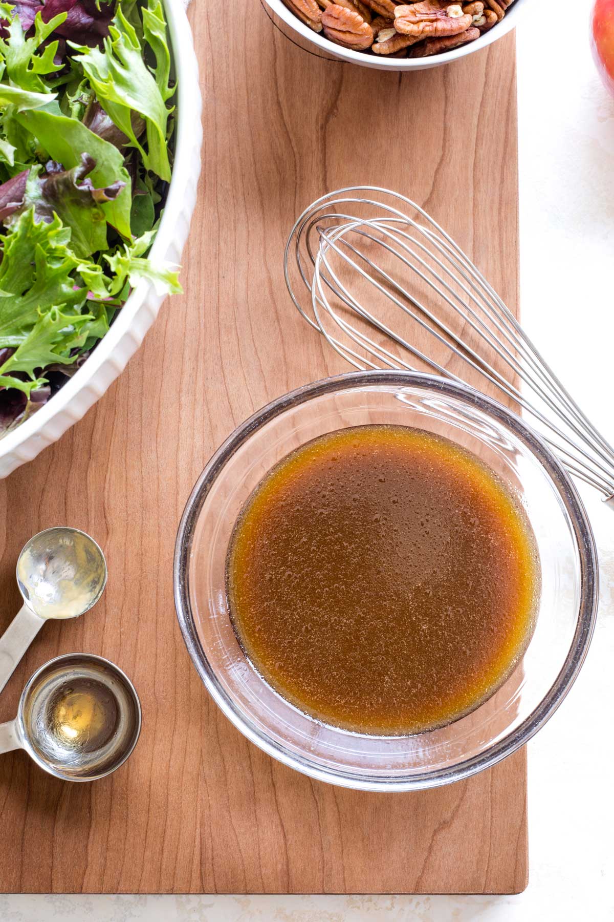 Small glass bowl of Balsamic Dressing with whisk, measuring spoons, bowls of salad and pecans nearby.
