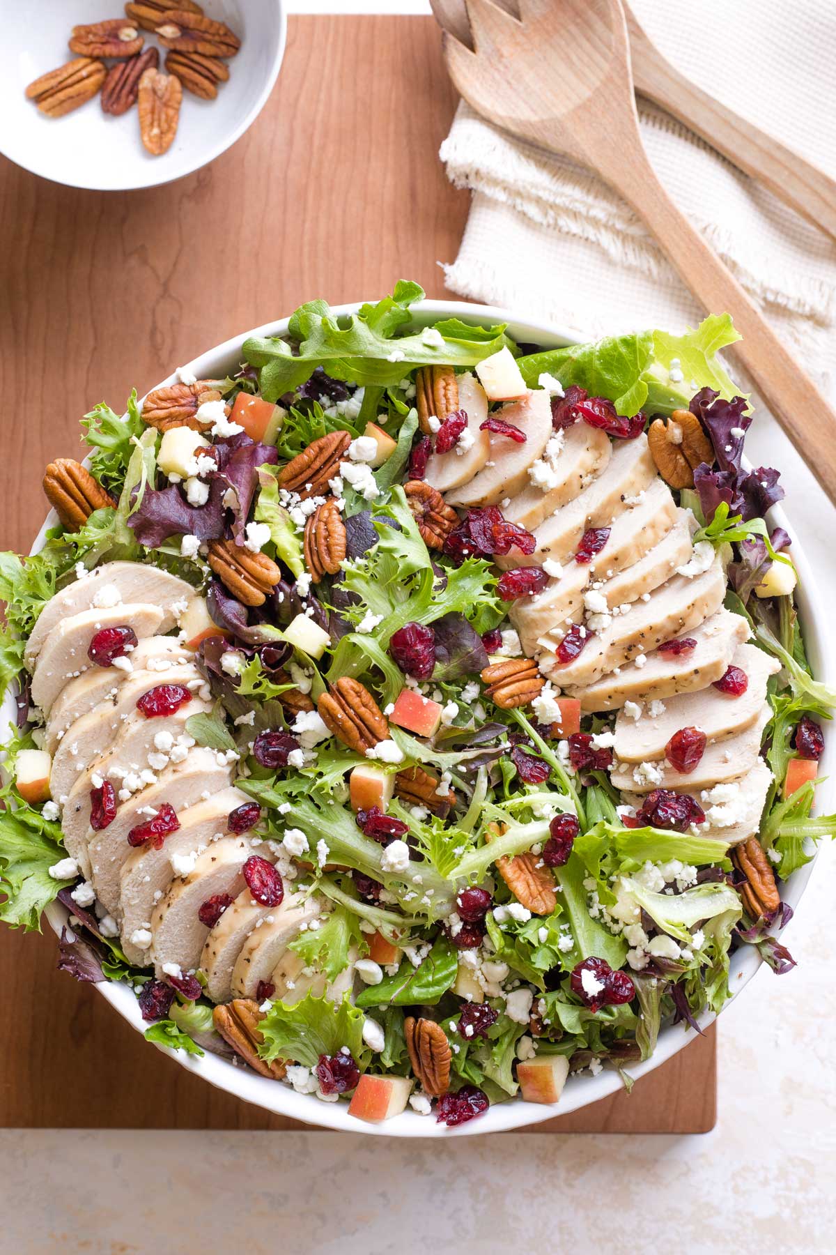 Overhead of salad arranged in serving bowl, with two lines of chicken fanned across; bowl of extra pecans nearby.