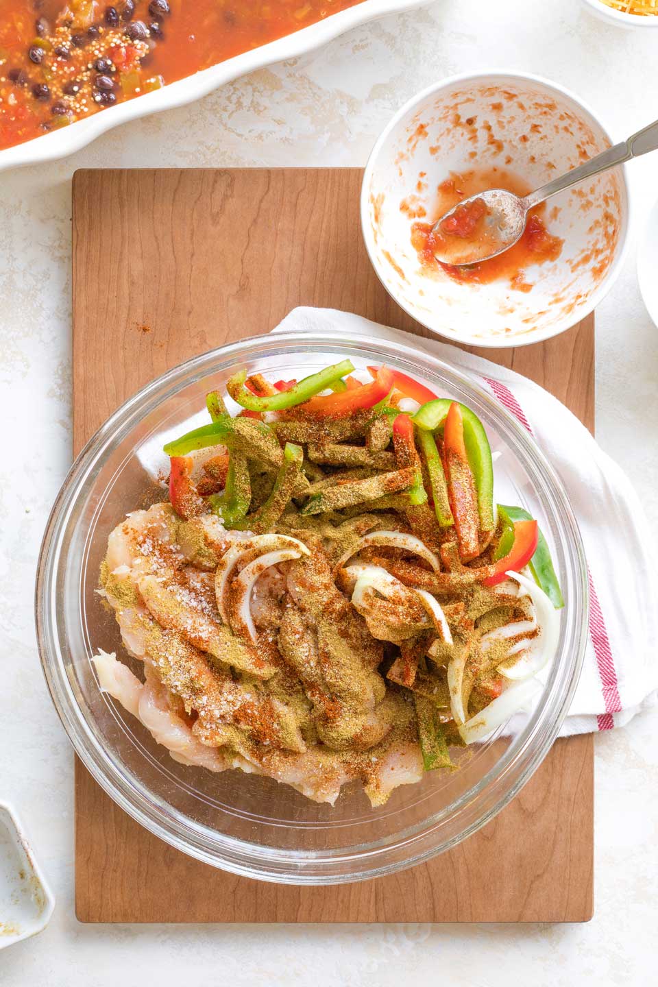 Overhead of fajita strips of chicken and vegetables in glass bowl, sprinkled with Mexican spices; casserole dish behind.