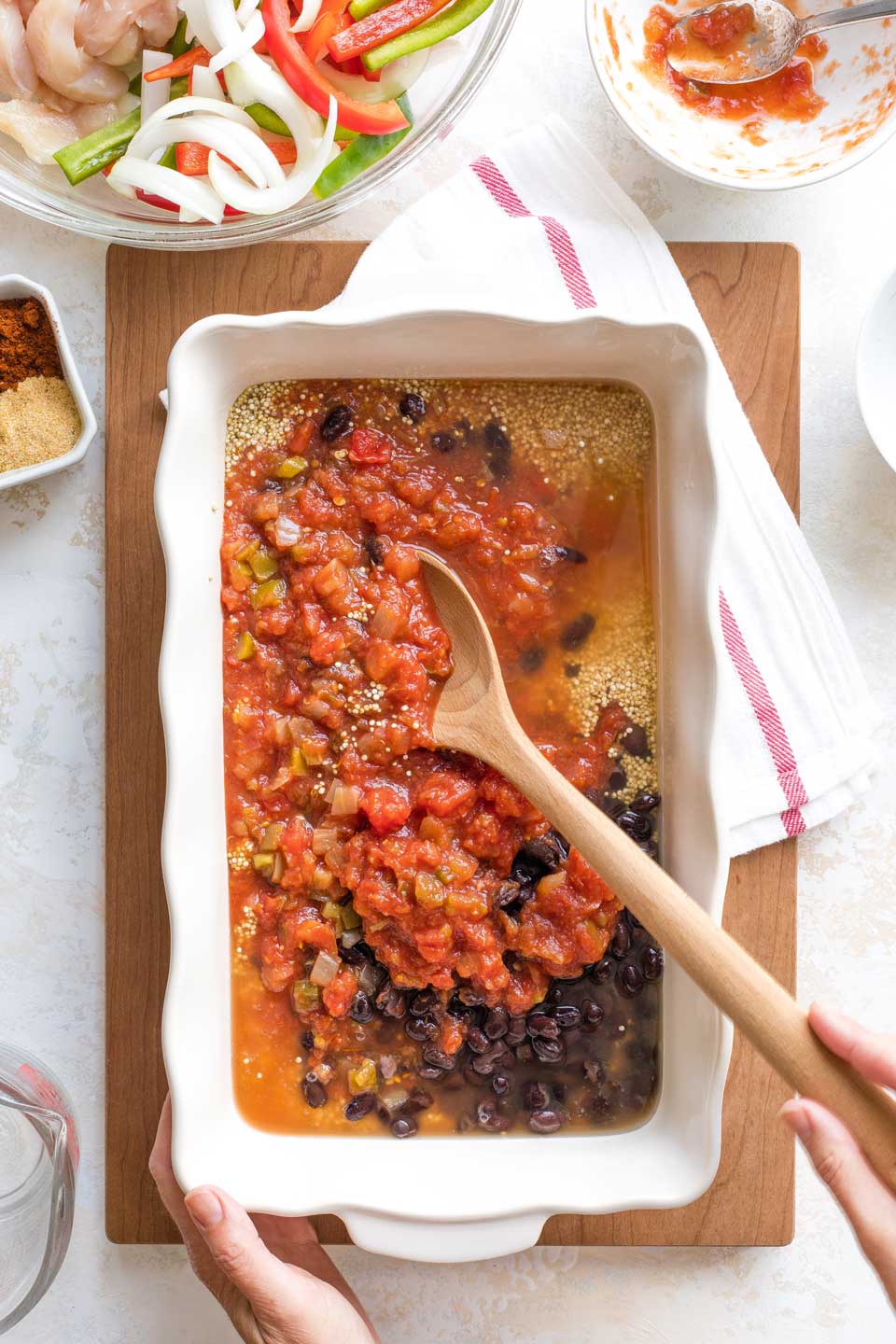 Hands holding casserole dish and wooden spoon, mixing Mexican black beans, salsa and quinoa base.
