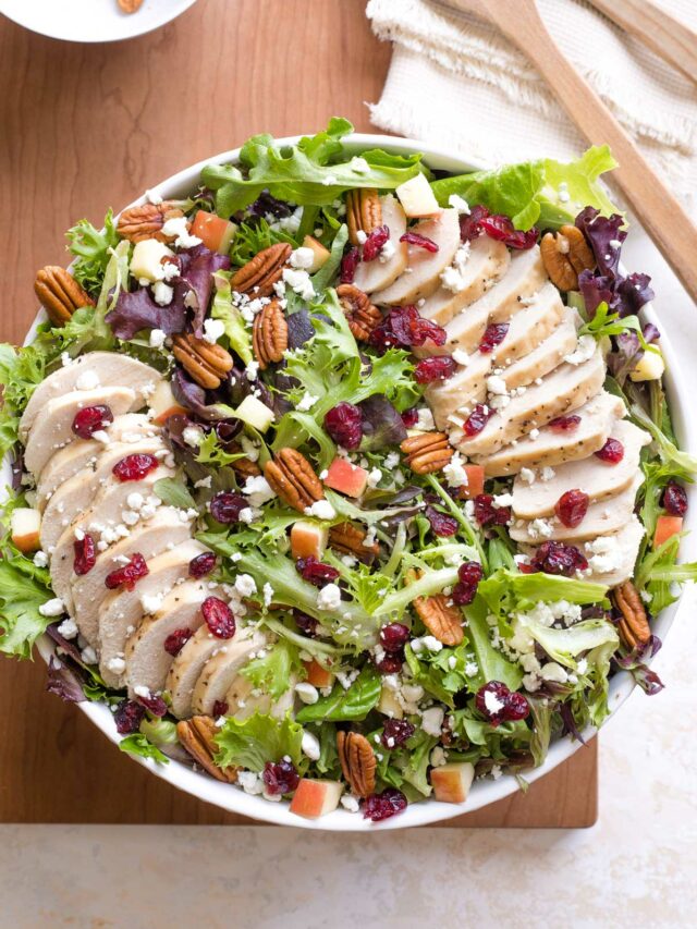 Overhead of salad arranged in serving bowl, with two lines of chicken fanned across; bowl of extra pecans nearby.