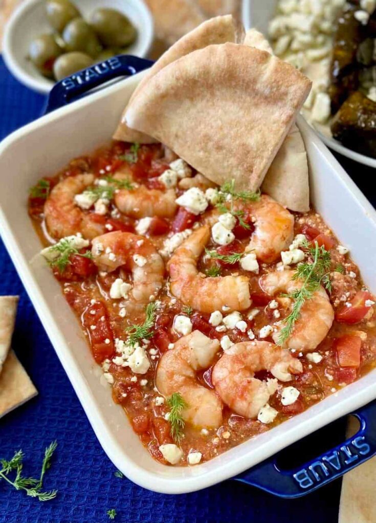 Baked dip in blue-handled baking dish with pita wedges and dishes of olives and feta.