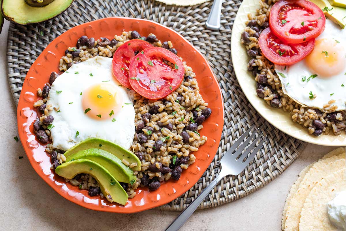 Two plates (bright orange and yellow) with servings of Gallo Pinto and toppings on woven mat with forks, tortilla stack and extra avocado nearby.