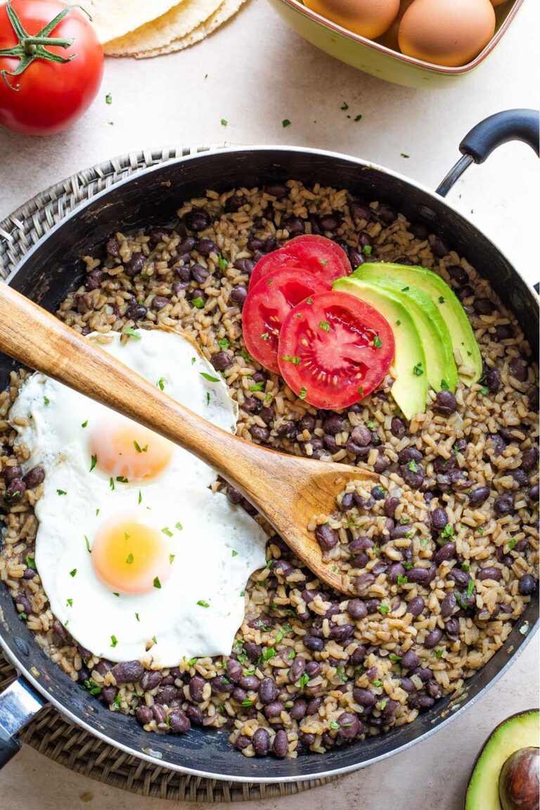 Overhead of cooked Gallo Pinto in pan with minced cilantro, avocado and tomato slices, 2 fried eggs on top.