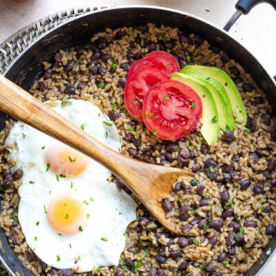 Flatlay of skillet full of cooked rice and beans, toppings added; extra tomato, avocado, eggs and tortillas nearby.