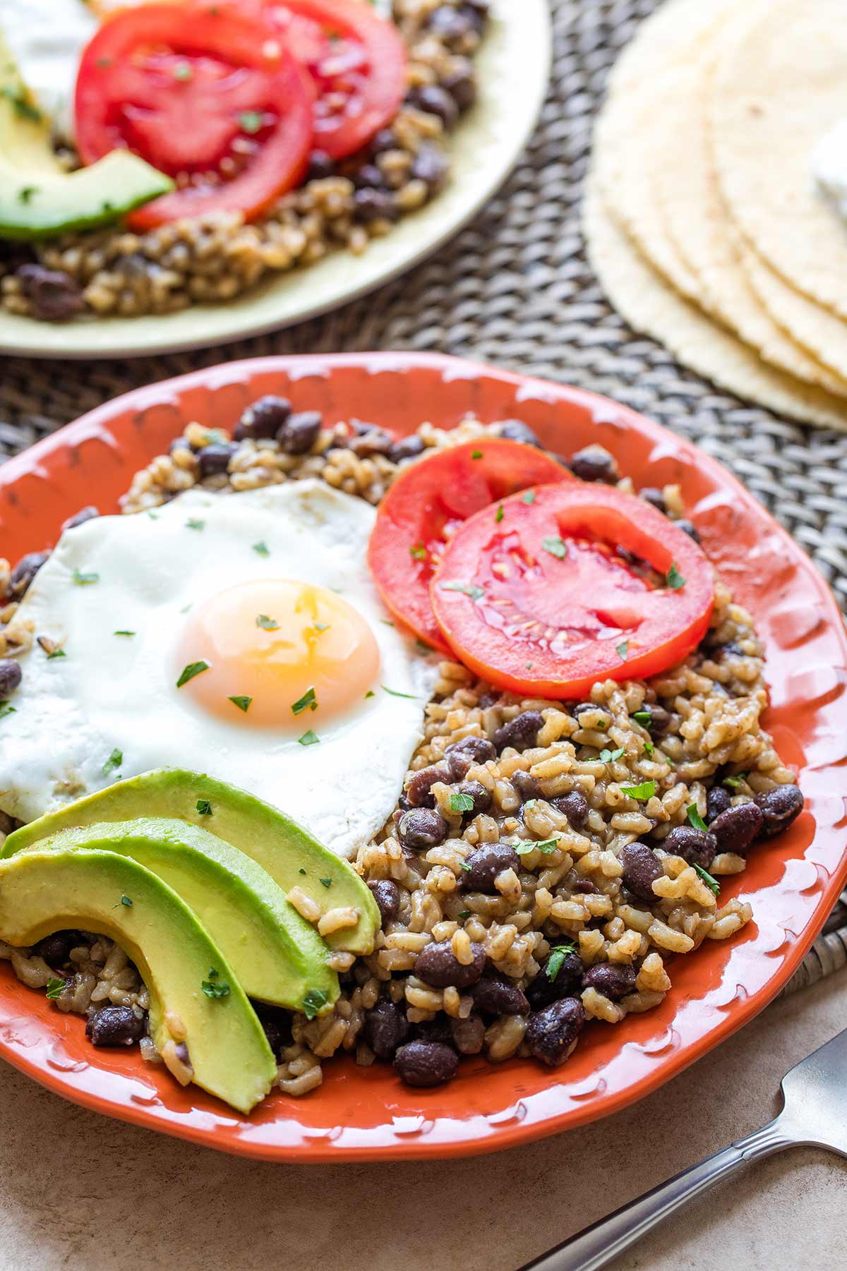 Rice and beans on orange plate with tomato, avocado and an egg nestled on top; second plateful in background.